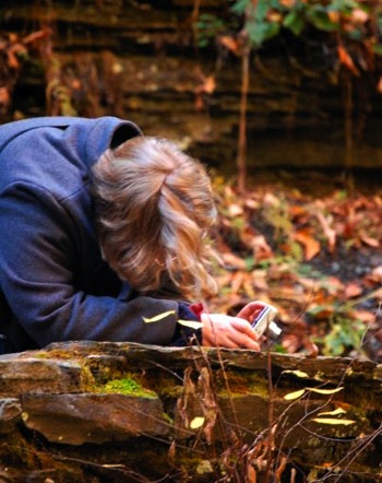 photograph of a fair-haired person wearing a gray coat and taking a photo in rocky/leafy background